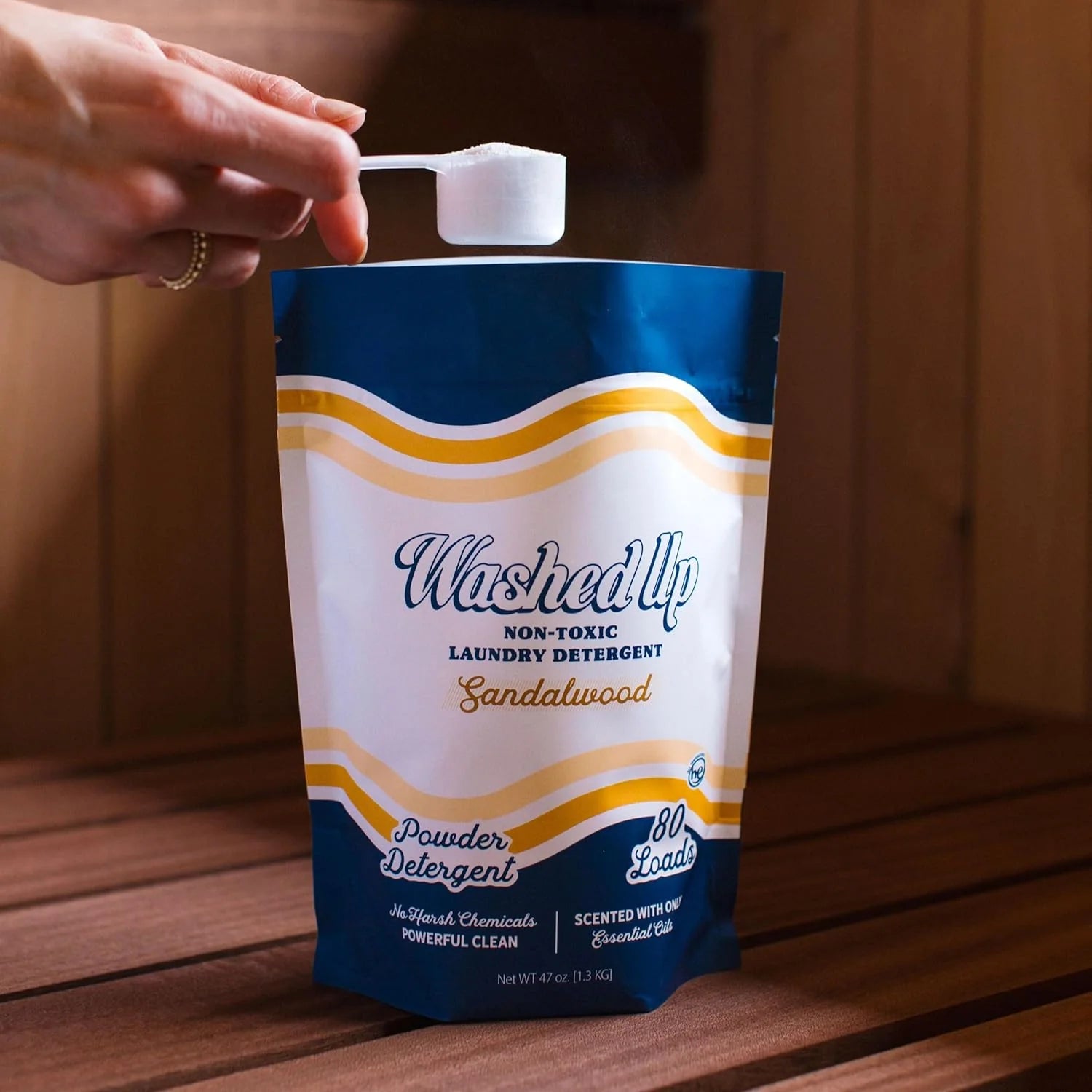 A hand holding a scoop above a pouch of "Washed Up" non-toxic sandelwood laundry detergent on a wooden surface, conveying cleanliness and eco-friendliness.