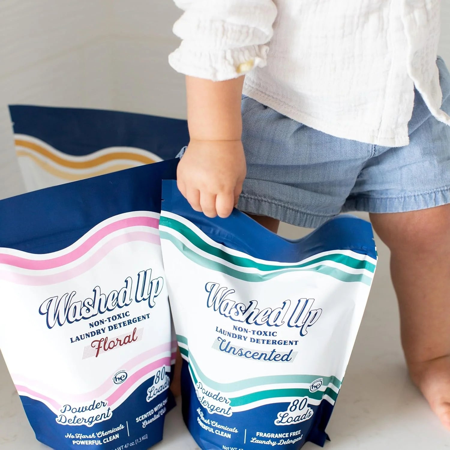 A toddler in blue shorts and a white shirt holds two colorful pouches of non-toxic laundry detergent labeled "Floral" and "Unscented." The scene feels fresh and clean.