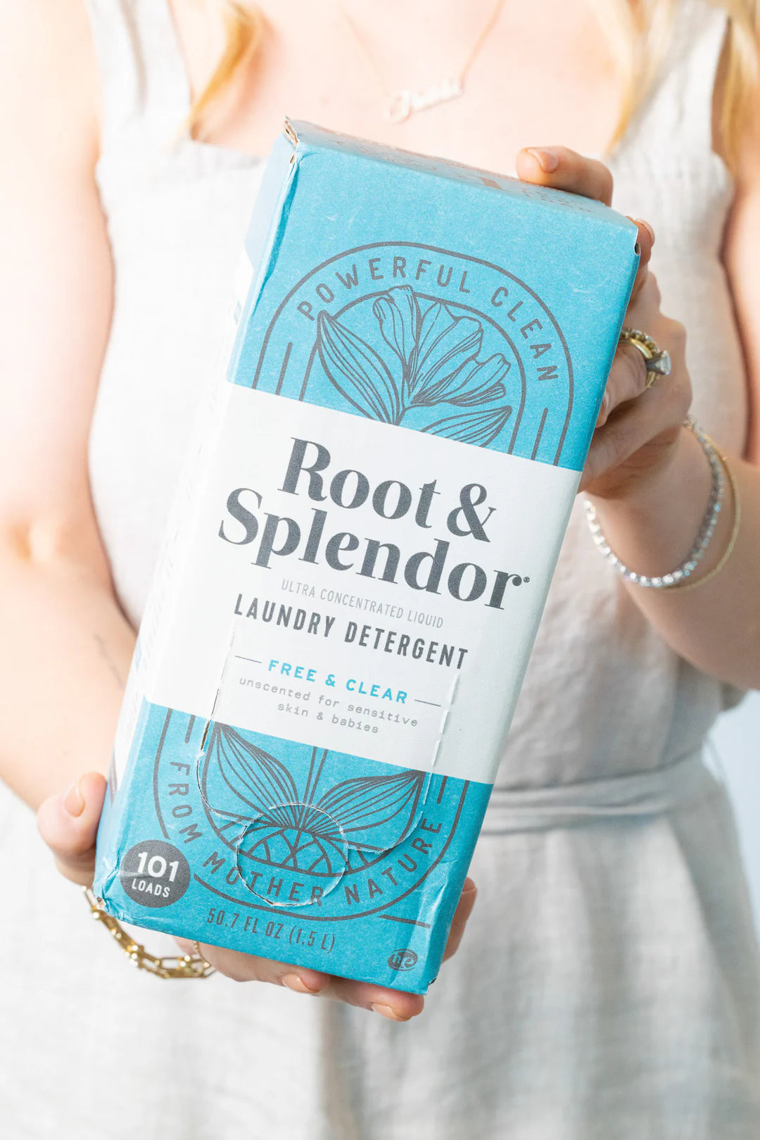 A woman in a light dress holds a blue and white box of "Root & Splendor" laundry detergent. The packaging highlights natural, powerful cleaning.
